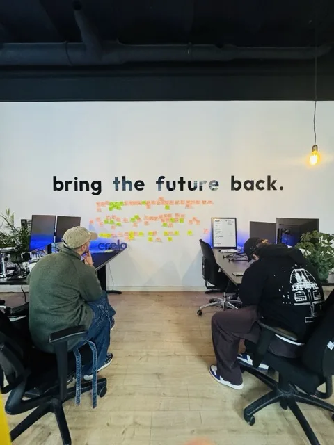 A studio wall reading bring the future back, covered in post-its, two people working in front of it