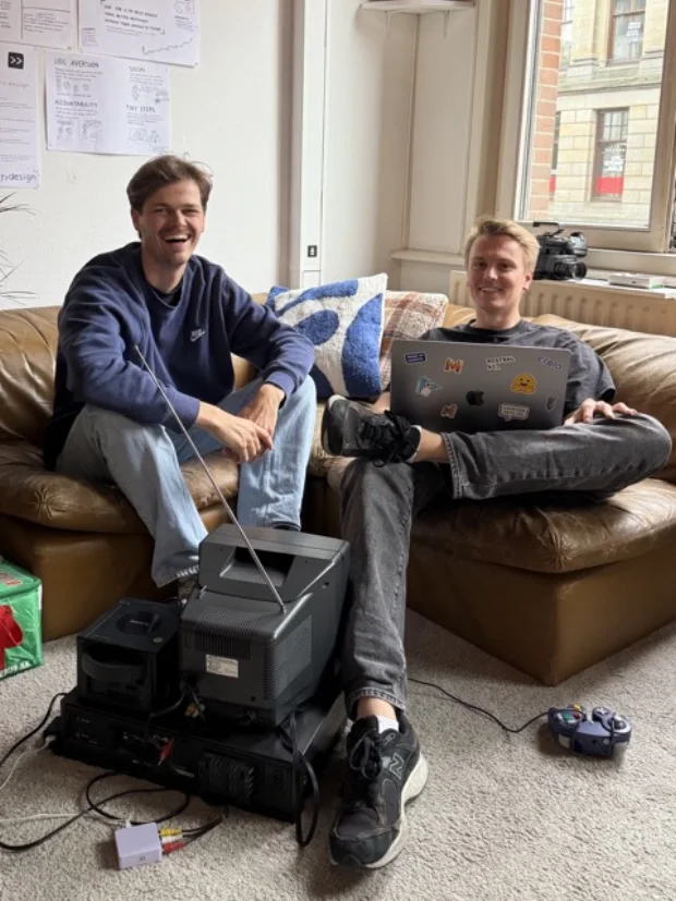 Rick and Robbin on a sofa in the studio, laughing, with an old CRT television and a games console on the floor